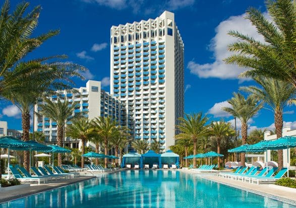 High-rise hotel with a geometric architectural design and numerous balconies, framed by tall palm trees. In the foreground, a large pool with turquoise lounge chairs and umbrellas lines either side. The sky is clear with a few scattered clouds.