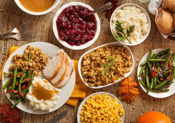 A Thanksgiving meal is spread out on a table with dishes including sliced turkey, mashed potatoes with gravy, stuffing, green beans, corn, cranberry sauce, dinner rolls, and a small pumpkin for decoration. The table is set with utensils and fall-themed accents.