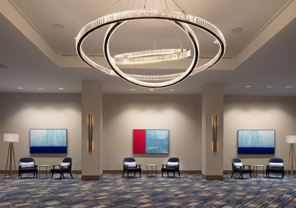 A modern lobby with a large circular chandelier hanging from the ceiling, illuminating the space. Six blue chairs and three small tables are arranged in front of three abstract paintings on the wall. Two floor lamps stand at either end of the seating area.