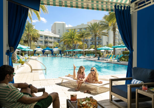 A man sits in a shaded cabana, looking at a woman and a girl lounging on poolside chairs by a large swimming pool. The pool area, surrounded by palm trees and sun umbrellas, features resort buildings in the background. The atmosphere is sunny and relaxed.