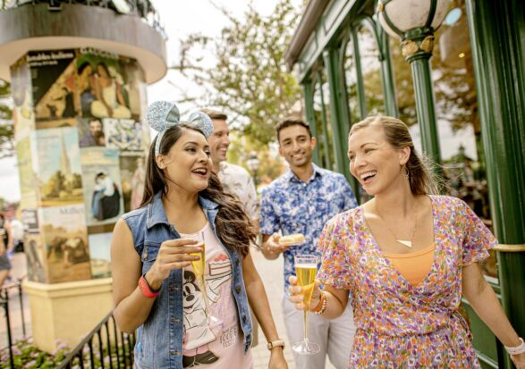 Four adults walk together outdoors at a lively park, smiling and holding drinks. The group appears happy, and the background features posters, greenery, and a building with green trim.