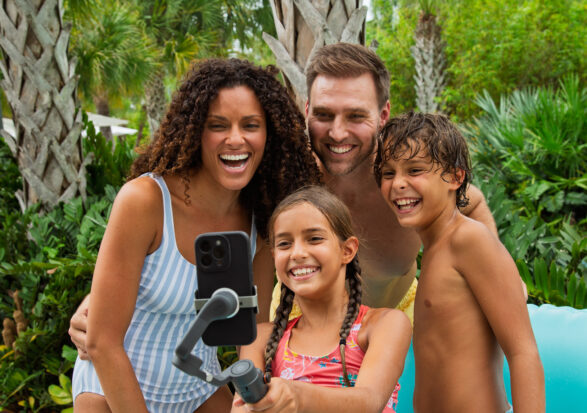A smiling family of four in swimsuits takes a selfie with a smartphone on a selfie stick, standing outdoors by palm trees on a sunny day.