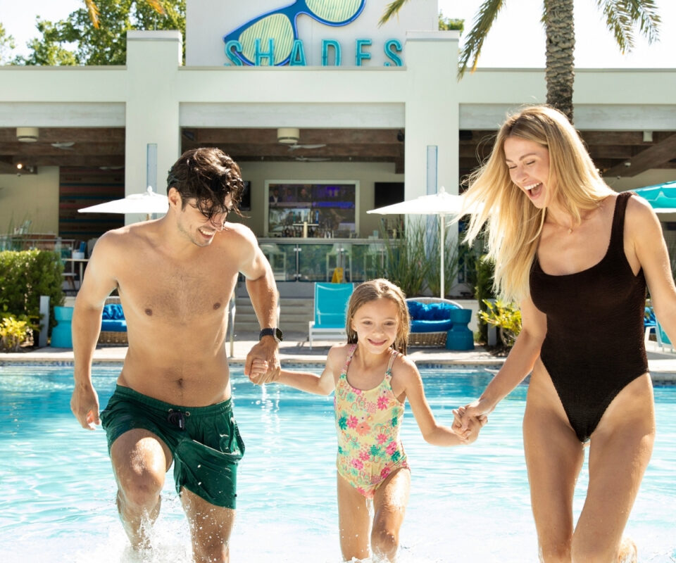 A man, woman, and young girl in swimsuits smile as they hold hands and walk out of a sunny swimming pool, with lounge chairs, palm trees, and a building labeled 